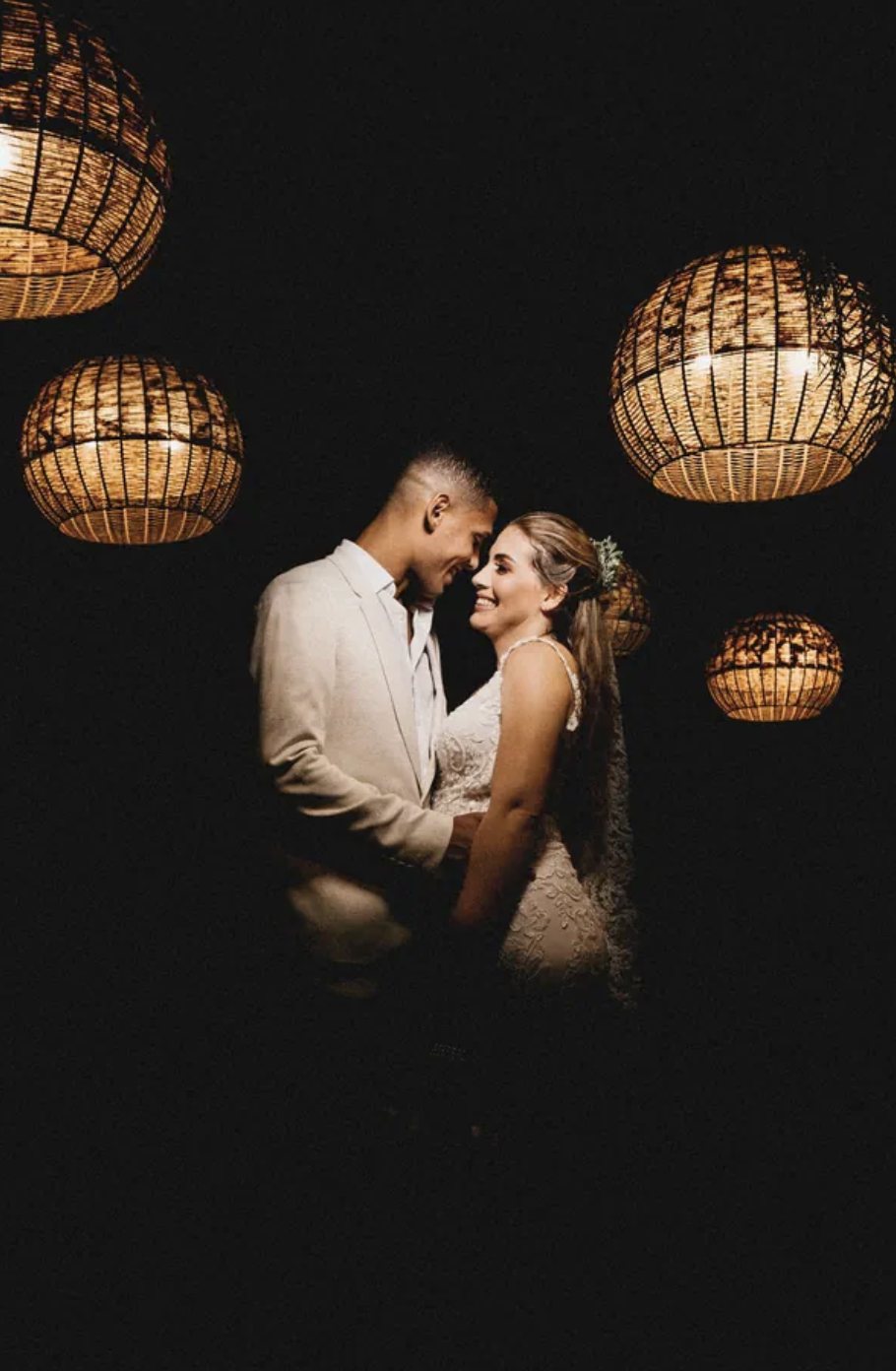 A bride and groom smiling forehead to forehead at night, surrounded by glowing woven pendant lanterns