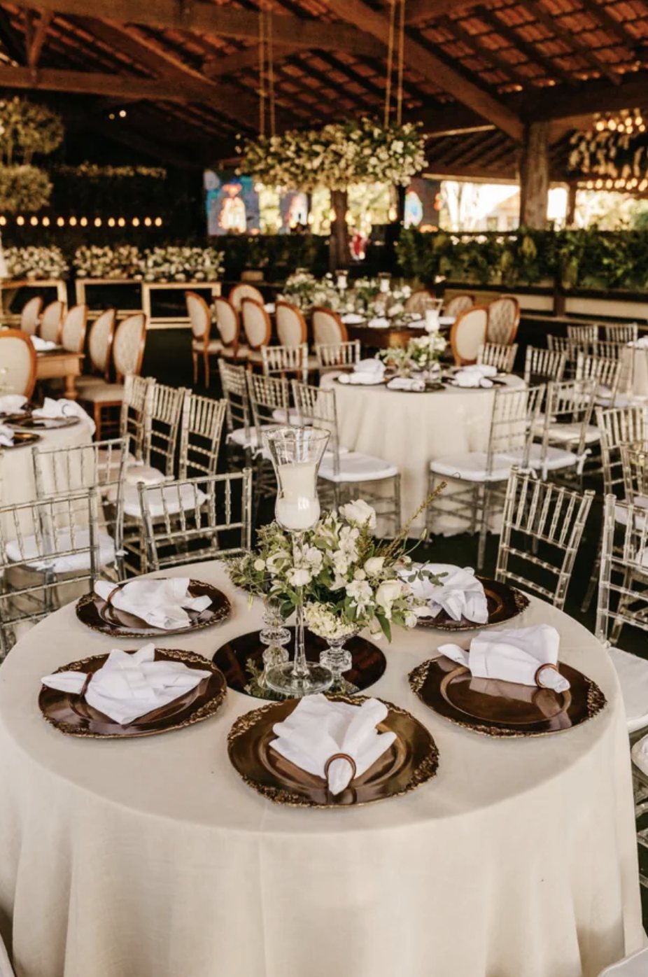 A round reception table set with gold-edged chargers, white linen napkins, and a centerpiece of white roses and greenery beneath a candle hurricane